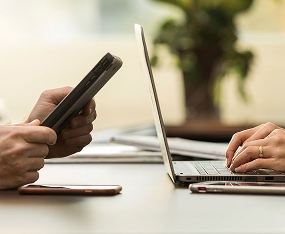 A pair of hands looking at a tablet while another pair types at a laptop