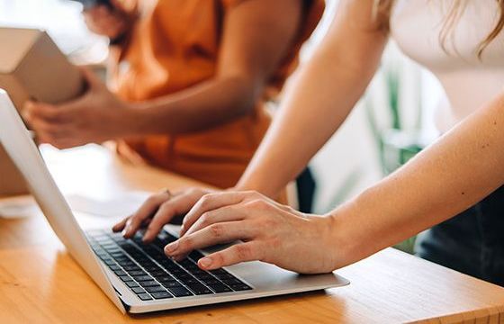 A woman working on a laptop while another person scans packages next to her