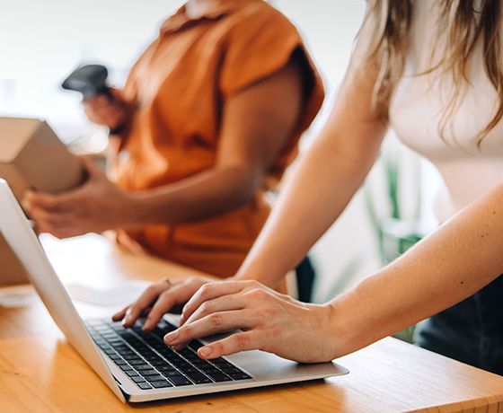 A woman working on a laptop while another person scans packages next to her